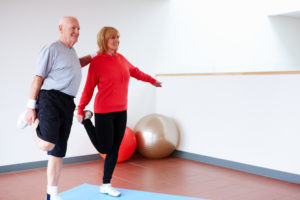 Senior man holds onto his wife for support as they both balance on one leg at the gym.